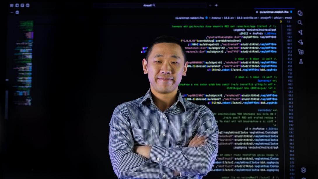 An individual in business attire stands with arms crossed in front of a wall of computer code